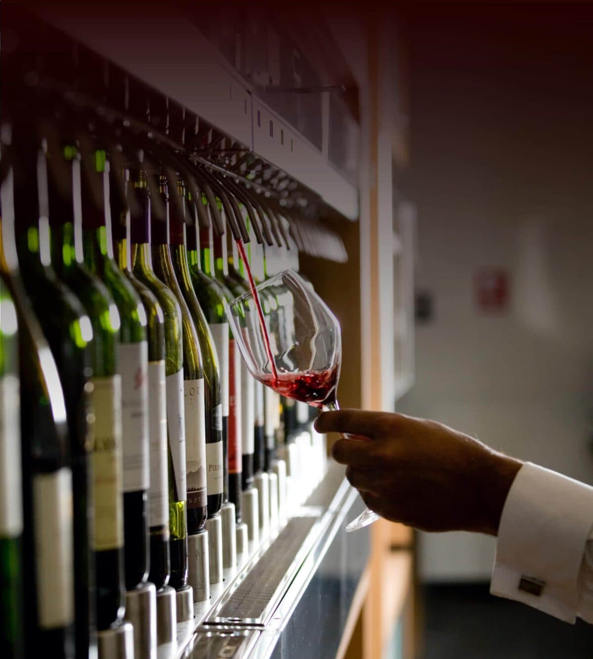 Hand tappar upp rött vin i ett glas från en Enomatic vinserveringsmaskin med flera vinflaskor i rad.