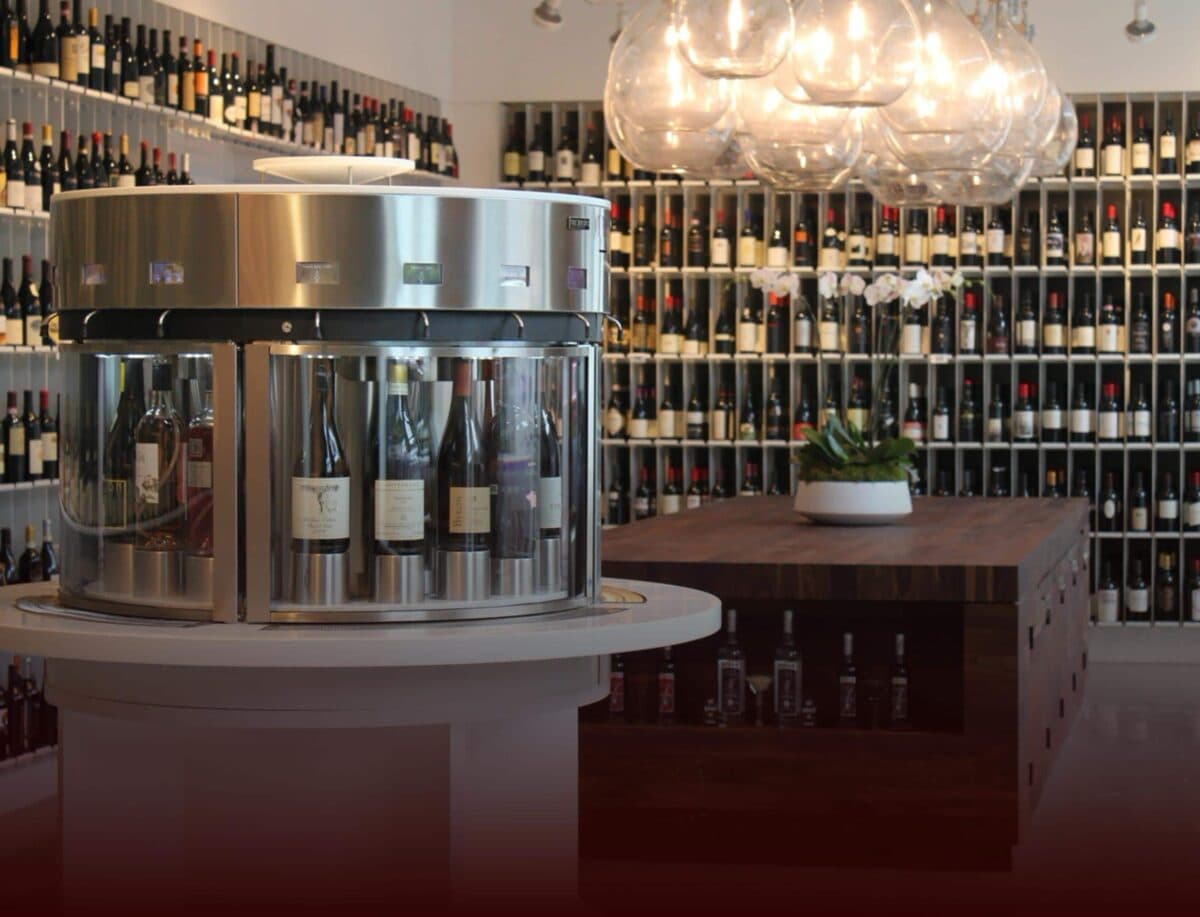 En rund Enomatic vinserveringsmaskin i en elegant vinbutik med väggar fyllda av vinflaskor och en rustik träbänk i bakgrunden.