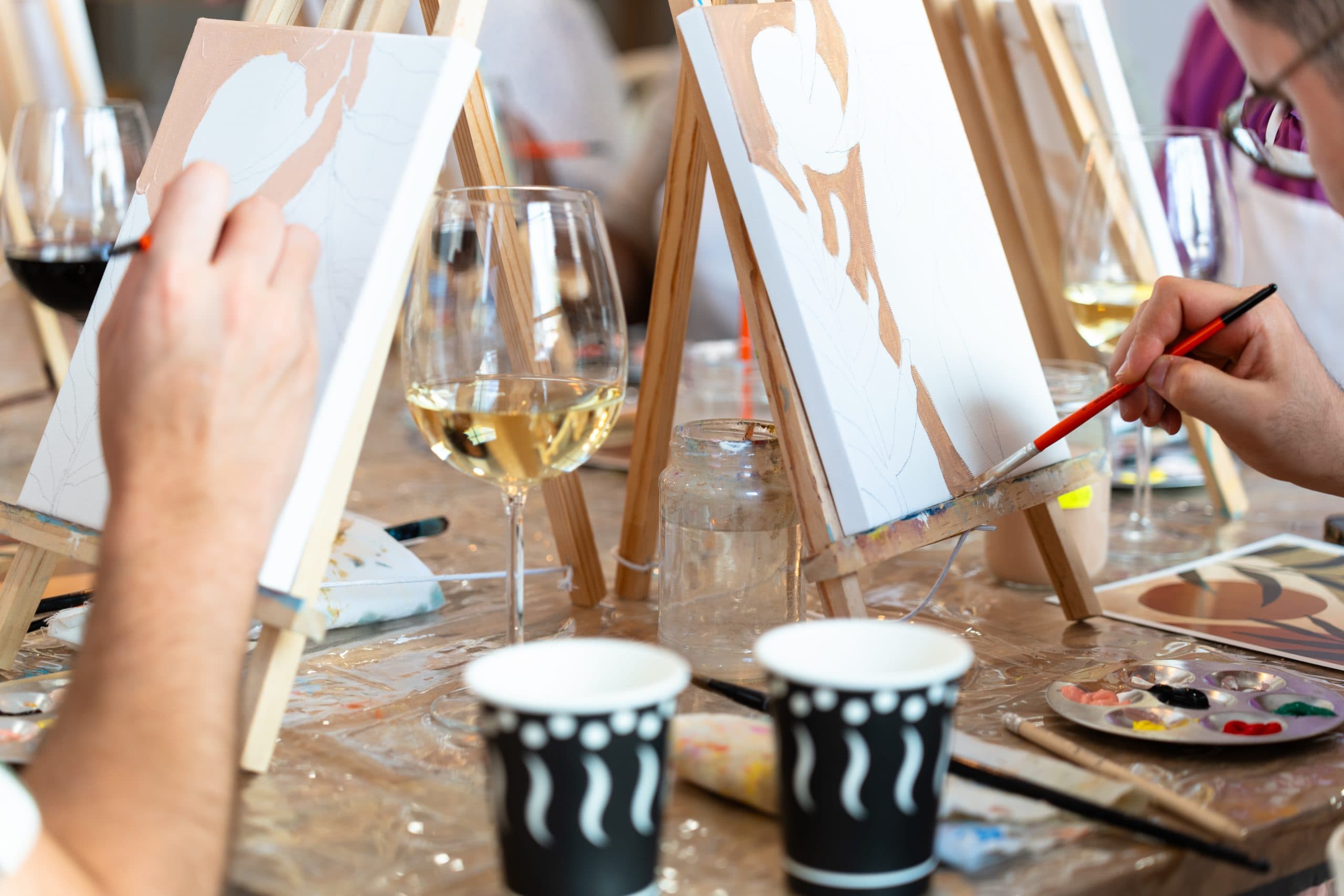 Två personer målar på canvas vid ett "sip and paint"-evenemang med vin och konstmaterial på bordet.